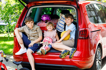 Three children, two boys and preschool girl sitting in car trunk before leaving for summer vacation with parents. Happy kids, siblings, brothers and sister with suitcases and toys going on journey