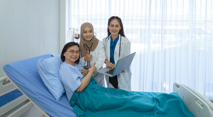 Muslim female doctor and young asian female doctor hold laptop with happy senior patient lie down on bed at hospital. Medicine and health care concept.