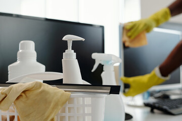 Group of detergents in white plastic bottles with dispensers and duster in basket against computer screen and gloved hands of man