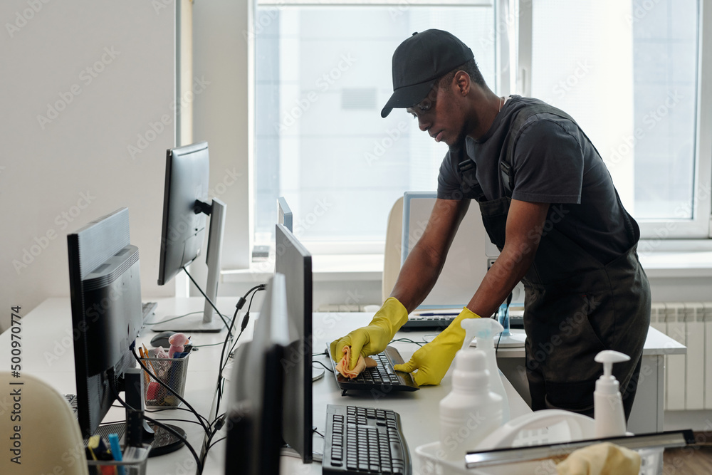 Poster Young contemporary black man in uniform cleaning keyboard of computer with duster and detergent in openspace office