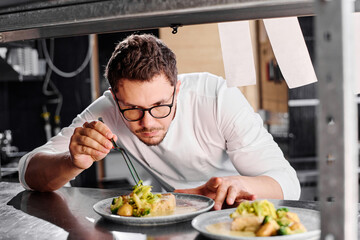 Young serious chef in eyeglasses decorating main dish on plate on counter before delivery