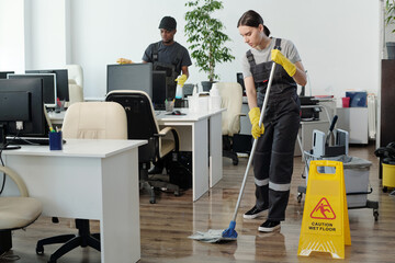 Young black man wiping computer monitors while Caucasian woman in coveralls and yellow gloves with mop cleaning floor in office