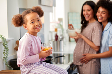 Pregnant Family With Two Mums Wearing Pyjamas Making Morning Pancakes In Kitchen With Daughter