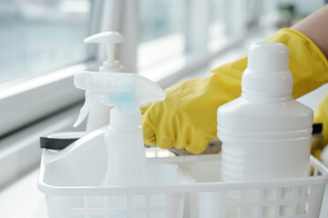 Group of white plastic bottles with detergents for washing windows in container against hands of female cleaner in yellow gloves