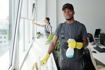 Young black man in coveralls, t-shirt, cap and rubber gloves using detergent in plastic sprayer while standing by window in office - obrazy, fototapety, plakaty