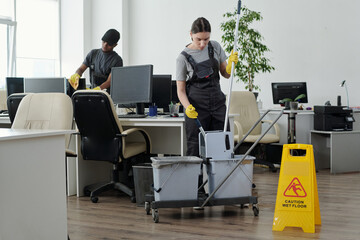 Young cleaning lady sqeezing mop in bucket against male colleague while going to wash floor in large modern openspace office