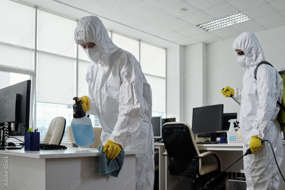 Poster workers of cleaning service in hazmat suits disinfecting furniture and computer equipment in large m