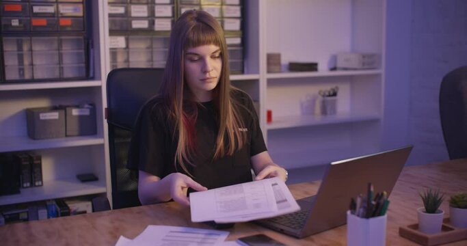 A young woman sits in the office and prints something into a laptop, then looks at her watch, someone bring documents to her and she continues to type, then she takes her phone. Slow motion 4k footage