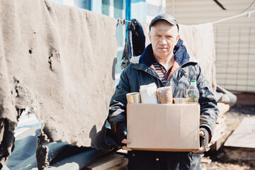 Concept humanitarian aid to refugees. Old man senior is holding cardboard box of donations canned food, looking camera