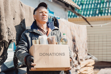 Old man senior is holding cardboard box of donations canned food, looking camera. Concept...