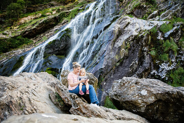 Cute toddler girl and mother sitting near water cascade of Powerscourt Waterfall, the highest...