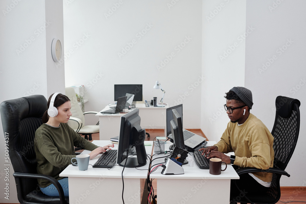 Poster Side view of two young intercultural programmers working in front of computers while decoding data or developing new software