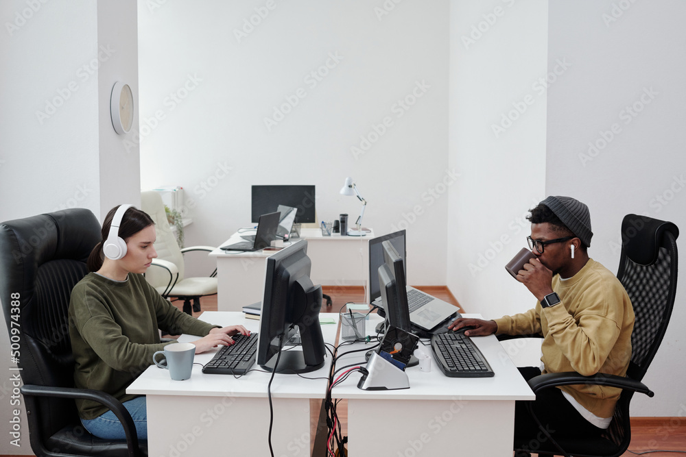 Poster Young male programmer having coffee and developing new program while sitting in front of computer and his female colleague in office
