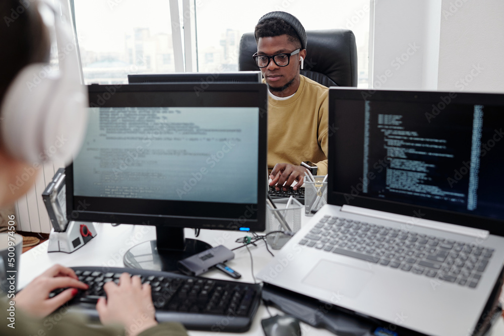 Poster Young serious male programmer looking at computer screen during network while sitting in front of his female colleague in office