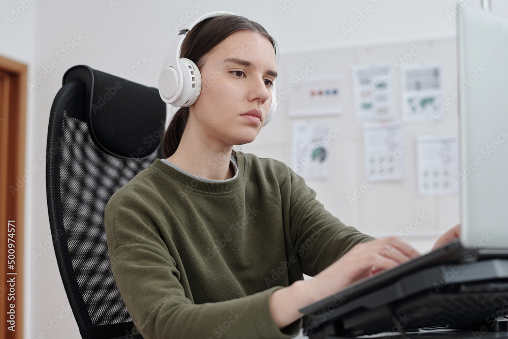 Poster Young serious female programmer in headphones and casualwear decoding data in front of computer monitor by her workplace