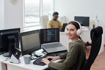 Happy young female it-engineer in headphones sitting by desk in front of computer monitor and...