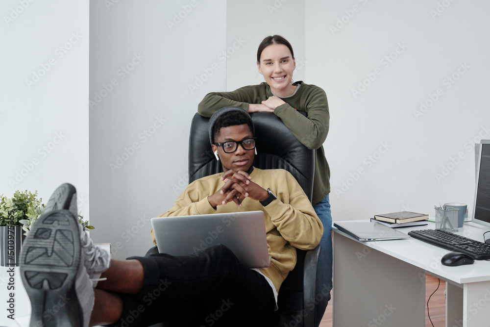 Poster young intercultural programmers in casualwear looking at camera by workplace, one of them smiling wh
