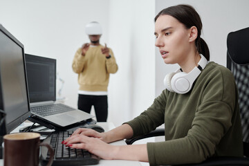 Young serious female programmer in casualwear decoding data and looking at computer screen against male coworker in vr headset