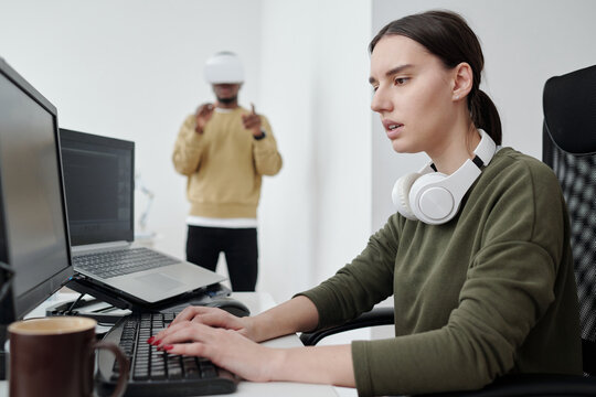 Young serious female programmer in casualwear decoding data and looking at computer screen against male coworker in vr headset - Powered by Adobe