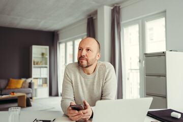 middle aged man in home office sitting in front of his laptop and holding a cell phone in his hand