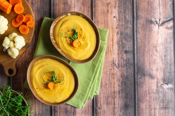 Vegan carrot and cauliflower puree soup garnished with green sprouts in ceramic bowls on a wooden table background with space for text. View from above. Banner