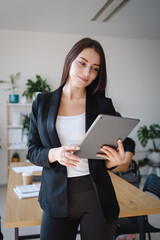 Young caucasian woman in formal suit stands indoors in the office with tablet in hands