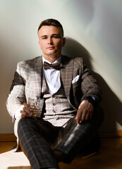 Portrait of thoughtful and handsome groom, wearing in brown plaid suit with bow tie, sitting on floor with glass of alcohol beverage on white background, seriously looking at camera