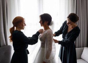 Side view of cheerful and pretty bride, standing near window in room, feeling happy while her friends correcting and tying her dress in wedding day - obrazy, fototapety, plakaty