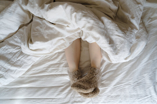 Feet in warm beige socks stick out of a blanket on a bed with white linens
