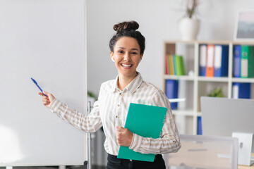 Presentation concept. Happy female manager holding notepad and pointing at free space on empty whiteboard in office
