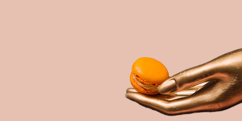 A golden female hand holds an orange macaroon, on a dirty pink background