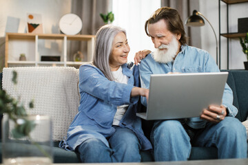 Happy mature family in casual outfit sitting together on couch and using wireless laptop. Smiling senior wife and husband spending time for communicating online with children and relatives at home