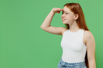 Little redhead kid smiling happy girl 12-13 years old in white tank shirt hold hand at forehead look far away distance isolated on plain green background studio portrait. Childhood lifestyle concept.