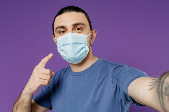 Young Man In Basic Blue T-shirt Sterile Face Mask Ppe To Safe From Coronavirus Virus Covid-19 Flu On Lockdown Quarantine Do Selfie Shot On Mobile Cell Phone Isolated On Plain Purple Color Background.