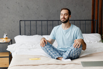 Young peaceful man in casual blue t-shirt sitting on bed in lotus pose yoga om aum gesture relax meditate try to calm down listen music in earphones rest relax spend time in bedroom home in own room.