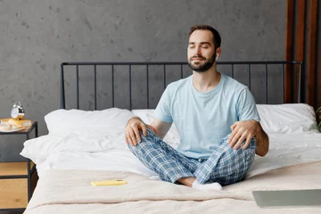 Foto op Plexiglas Young peaceful man in casual blue t-shirt sitting on bed in lotus pose yoga om aum gesture relax meditate try to calm down listen music in earphones rest relax spend time in bedroom home in own room. © ViDi Studio