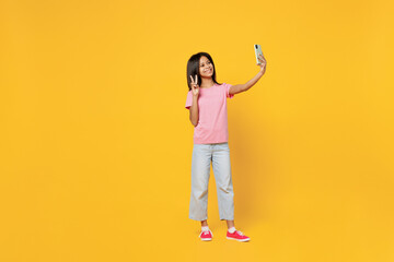 Fototapeta na wymiar Full size little kid girl of African American ethnicity 12-13 years old in pink t-shirt do selfie shot on mobile cell phone show v-sign isolated on plain yellow background. Childhood lifestyle concept