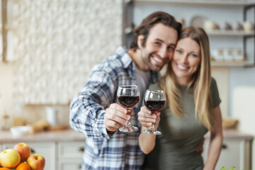 Glad millennial european couple hugs, clinking glasses of wine and look at camera at scandinavian kitchen interior