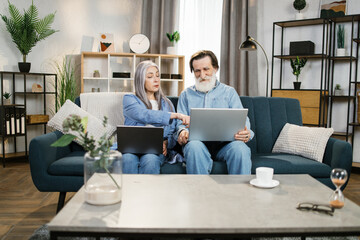 Mature man and woman using laptops, looking at screen, older spouses browsing apps, shopping or chatting online, making video call, using bank service, sitting on cozy sofa in living room