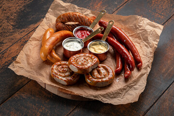 A mix of grilled sausages - Bavarian, round, Cumberland, bratwurst with ketchup and sauce on a wooden table. Traditional Bavarian Beer Snack