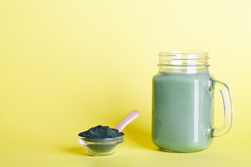 Obraz na płótnie Canvas fresh green smoothie in glass bottle and spirulina powder with spoon on yellow background. useful habits, self care and healthy lifestyle. copy space