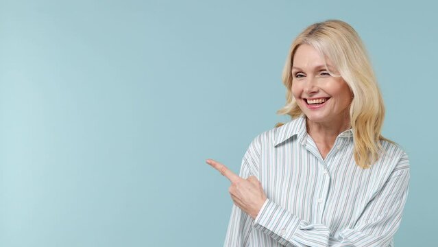 Promoter elderly blonde woman lady 40s years old in white shirt point finger aside on workspace copy space mockup promo commercial area isolated on plain pastel light blue background studio portrait