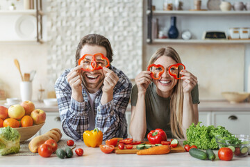 Glad funny young european couple cooking at table with organic vegetables, have fun and put pieces of pepper to eyes