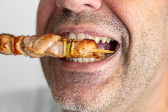 Mouth of a middle-aged man biting into a skewer of meat.