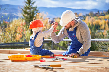 Father with toddler son building wooden frame house. Male builder giving high five to kid on...