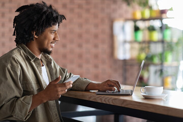 Happy young african american curly man with smartphone typing on laptop, sits at table with cup