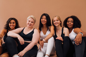 Five smiling women in fitness clothes sitting together on pastel background. Females with different...