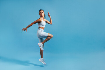 Fototapeta na wymiar Slim woman jumping in studio on blue background. Muscular female in fitness clothes exercising.