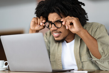 Concentrated young black curly guy in glasses squints and looks at laptop at table with cup in cafe...