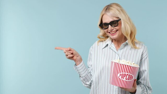 Elderly blonde woman lady 40s years old wears white shirt sunglasses hold bucket of popcorn point on workspace area copy space mock up isolated on plain pastel light blue background studio portrait